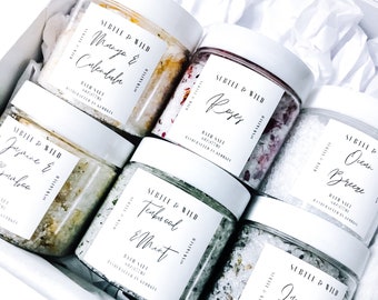 May include: A set of six bath salts in glass jars with white labels. The labels have black text and list the scents: Mango & Calendula, Roses, Jasmine & Bamboo, Teawood & Mint, Lavender, and Ocean Breeze. The jars are arranged in a white box with white tissue paper.