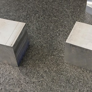 May include: Two silver metal blocks, each with a smooth, rectangular surface. The blocks are sitting on a gray, textured surface.