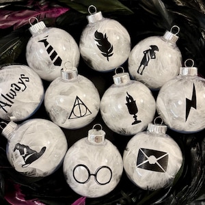 May include: A collection of clear glass Christmas ornaments filled with white feathers. Each ornament features a black graphic, including a lightning bolt, glasses, and the word "Always".