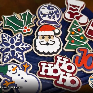 May include: A collection of 12 wooden Christmas ornaments featuring various holiday themes, including a snowman, Santa Claus, a Christmas tree, a snowflake, holly, a gift box, a stocking, a snow globe, and the words "Ho Ho Ho" and "Joy".