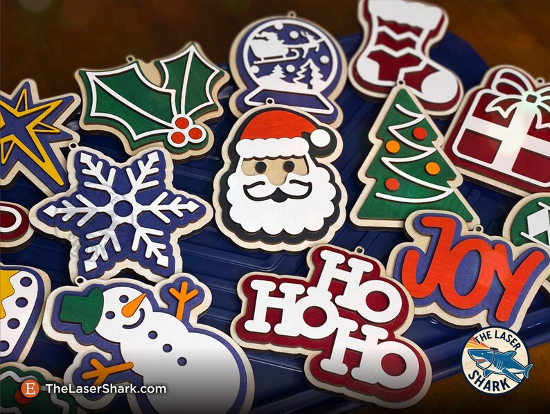May include: A collection of 12 wooden Christmas ornaments featuring various holiday themes, including a snowman, Santa Claus, a Christmas tree, a snowflake, holly, a gift box, a stocking, a snow globe, and the words "Ho Ho Ho" and "Joy".