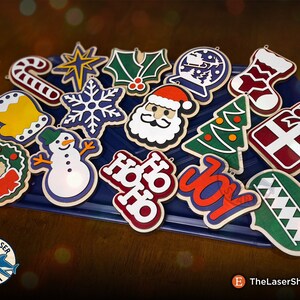 May include: A collection of 16 wooden Christmas ornaments in various shapes and colors, including a snowman, a star, a stocking, a Santa Claus, a Christmas tree, a wreath, a candy cane, a snowflake, a bell, a holly sprig, a gift box, a globe, and a "Ho Ho Ho" and "Joy" sign. The ornaments are arranged on a blue tray.