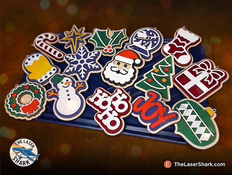 May include: A collection of 16 wooden Christmas ornaments in various shapes and colors, including a snowman, a star, a stocking, a Santa Claus, a Christmas tree, a wreath, a candy cane, a snowflake, a bell, a holly sprig, a gift box, a globe, and a "Ho Ho Ho" and "Joy" sign. The ornaments are arranged on a blue tray.