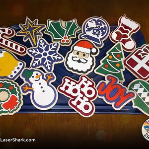 May include: A collection of 15 wooden Christmas ornaments with various designs, including a candy cane, a star, holly, a snowflake, Santa Claus, a Christmas tree, a gift box, a bell, a snowman, a wreath, the words "Ho Ho Ho", the word "Joy", and a green ornament with a diamond pattern.
