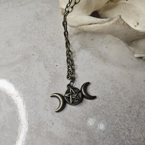 May include: Antique bronze necklace with a triple moon pendant and a central pentagram. The pendant hangs from a matching chain. The necklace is set against a neutral backdrop, emphasizing the intricate details of the celestial design.