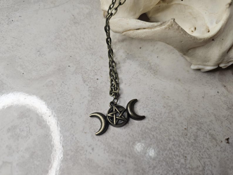 May include: Antique bronze necklace with a triple moon pendant and a central pentagram. The pendant hangs from a matching chain. The necklace is set against a neutral backdrop, emphasizing the intricate details of the celestial design.