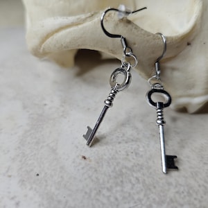 May include: A pair of silver key-shaped earrings with a small circle at the top of each key. The earrings are hanging from a white surface.