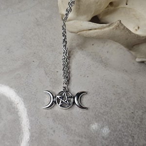 May include: A silver pendant necklace with a triple moon and pentagram design. The necklace hangs from a fine chain. The background is a light grey surface with a skull in the upper right corner.