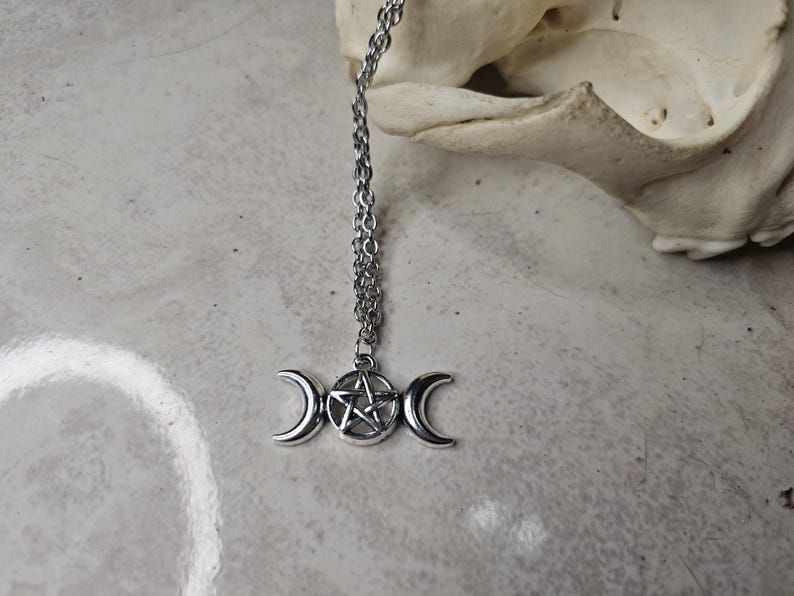 May include: A silver pendant necklace with a triple moon and pentagram design. The necklace hangs from a fine chain. The background is a light grey surface with a skull in the upper right corner.