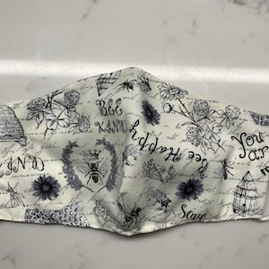 May include: A white face mask with a black and white bee and floral print. The mask has the text "Bee Kind" and "You Are My Sunshine" printed on it.