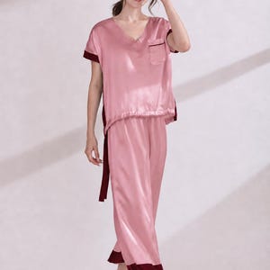 May include: A two-piece pajama set in light pink with dark maroon trim. The top features a V-neckline, short sleeves, and a single chest pocket. The matching pants are loose-fitting with a straight-leg cut, both pieces finished with dark maroon cuffs.