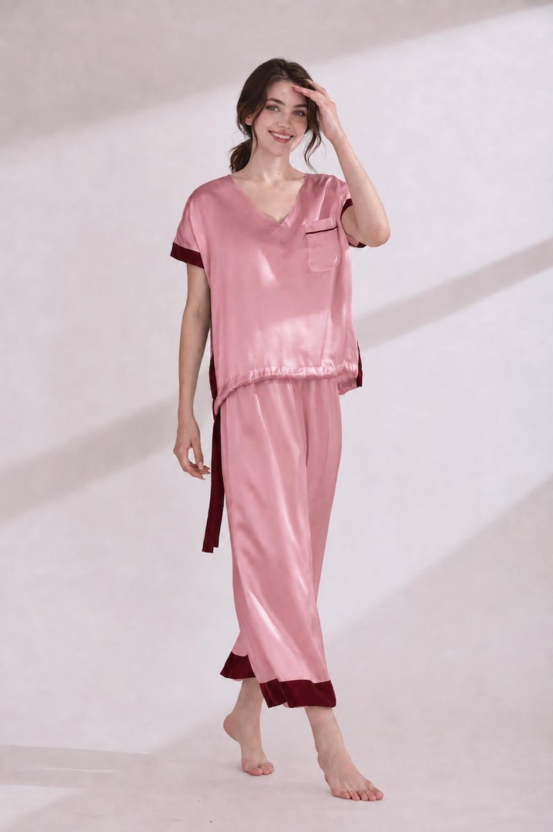 May include: A two-piece pajama set in light pink with dark maroon trim. The top features a V-neckline, short sleeves, and a single chest pocket. The matching pants are loose-fitting with a straight-leg cut, both pieces finished with dark maroon cuffs.