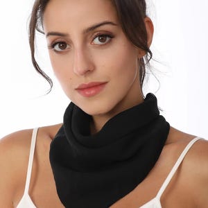 May include: A solid black square scarf tied around the neck in a triangular fold. The scarf is made of a lightweight, semi-sheer fabric that drapes softly. It is worn over a simple white vest top with thin shoulder straps against a plain background.