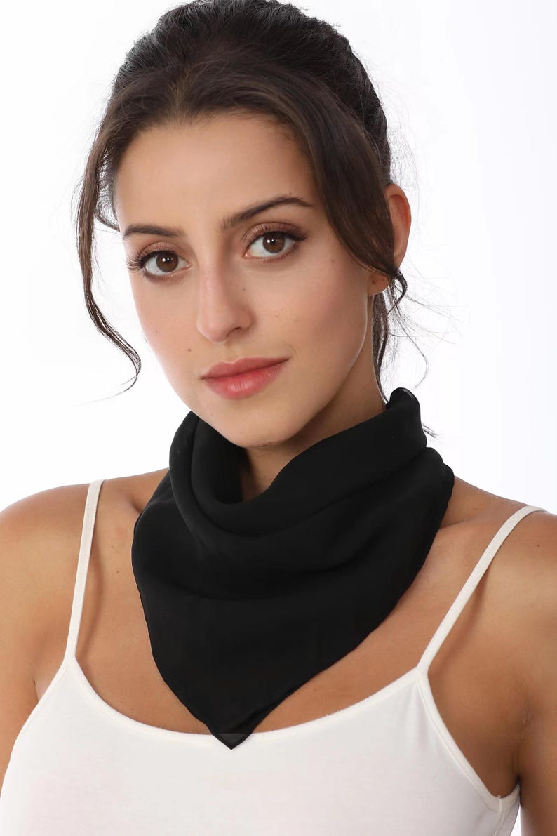 May include: A solid black square scarf tied around the neck in a triangular fold. The scarf is made of a lightweight, semi-sheer fabric that drapes softly. It is worn over a simple white vest top with thin shoulder straps against a plain background.