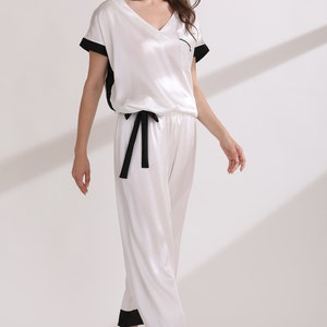 May include: A two-piece pajama set consisting of a white V-neck short-sleeve top with black trim on the sleeves and a chest pocket, paired with matching white cropped pants featuring a black drawstring waist and black cuffs at the hem.