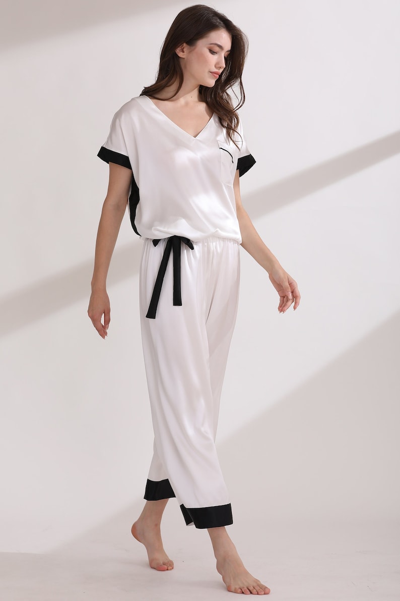 May include: A two-piece pajama set consisting of a white V-neck short-sleeve top with black trim on the sleeves and a chest pocket, paired with matching white cropped pants featuring a black drawstring waist and black cuffs at the hem.
