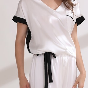 May include: A white V-neck top with short sleeves featuring black trim on the cuffs and sides, a chest pocket with black piping, and a matching white pair of pants with a black drawstring tie at the waist, all made from a smooth fabric.