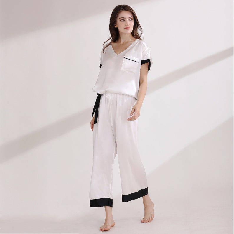 May include: A white two-piece pajama set featuring a short-sleeved V-neck top with a chest pocket and black trim, paired with matching wide-leg cropped trousers that have a black waistband tie and black hem detailing on the cuffs.