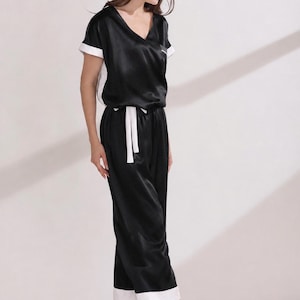 May include: A black pajama set featuring a short-sleeved top with a V-neckline and white trim on the sleeves and sides. The matching pants have a white drawstring waist and white cuffs at the hem, creating a coordinated two-piece loungewear outfit.