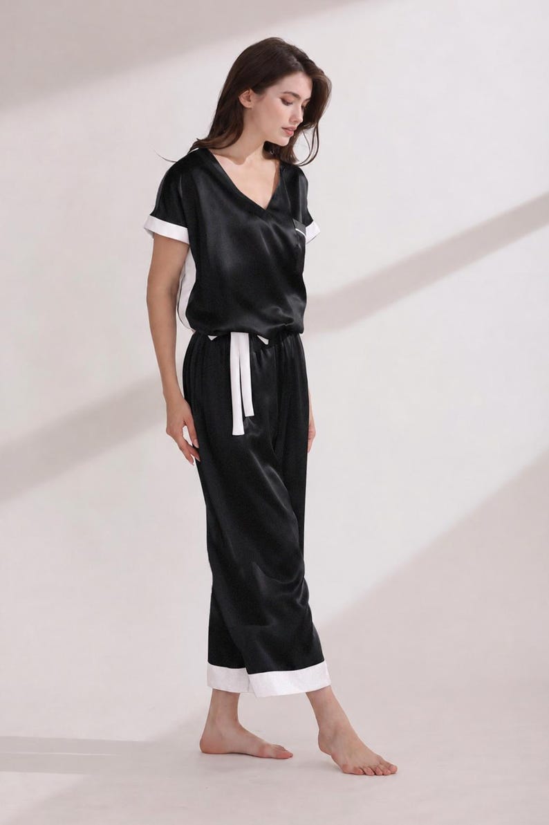 May include: A black pajama set featuring a short-sleeved top with a V-neckline and white trim on the sleeves and sides. The matching pants have a white drawstring waist and white cuffs at the hem, creating a coordinated two-piece loungewear outfit.