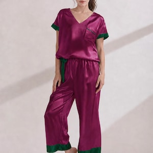 May include: A deep magenta satin pajama set featuring a short-sleeved V-neck top with a chest pocket and wide-leg cropped trousers. The set is accented with contrasting dark green trim on the sleeve cuffs, pocket edge, and trouser hems.