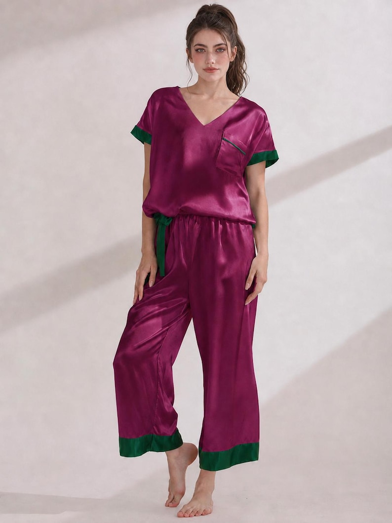 May include: A deep magenta satin pajama set featuring a short-sleeved V-neck top with a chest pocket and wide-leg cropped trousers. The set is accented with contrasting dark green trim on the sleeve cuffs, pocket edge, and trouser hems.