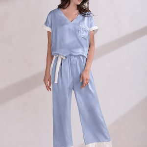 May include: A light blue pajama set consisting of a short-sleeved V-neck top with a chest pocket and wide-leg trousers. Both the top and the trousers feature contrasting white trim on the sleeve cuffs, pocket edge, and pant hems, with a white tie.