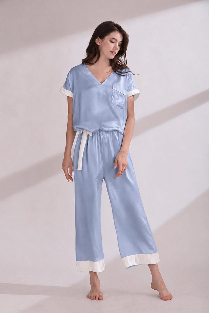 May include: A light blue pajama set consisting of a short-sleeved V-neck top with a chest pocket and wide-leg trousers. Both the top and the trousers feature contrasting white trim on the sleeve cuffs, pocket edge, and pant hems, with a white tie.