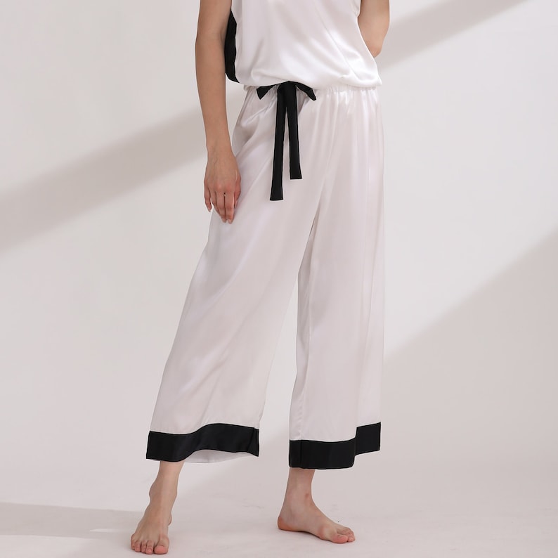 May include: A white satin pajama set consisting of a sleeveless top and wide-leg cropped trousers. The trousers feature a black drawstring waist and a thick black hem at the bottom. The top has black side panels and a smooth, lustrous finish.