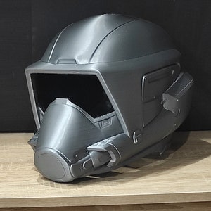 Op de afbeelding: Een grijze 3D-geprinte helm met een vizier en een gezichtsmasker. De helm heeft een futuristisch ontwerp met verschillende details en texturen.