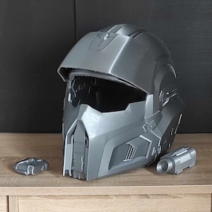 May include: A gray 3D printed helmet with a visor and a small gray accessory attached to the side. The helmet is sitting on a wooden surface.
