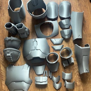 Puede incluir: Una colecci&oacute;n de piezas de armadura grises impresas en 3D, que incluyen petos, protectores de hombros y componentes de brazos. Las piezas est&aacute;n dispuestas sobre una superficie de madera, mostrando las diversas partes de un disfraz o accesorio.