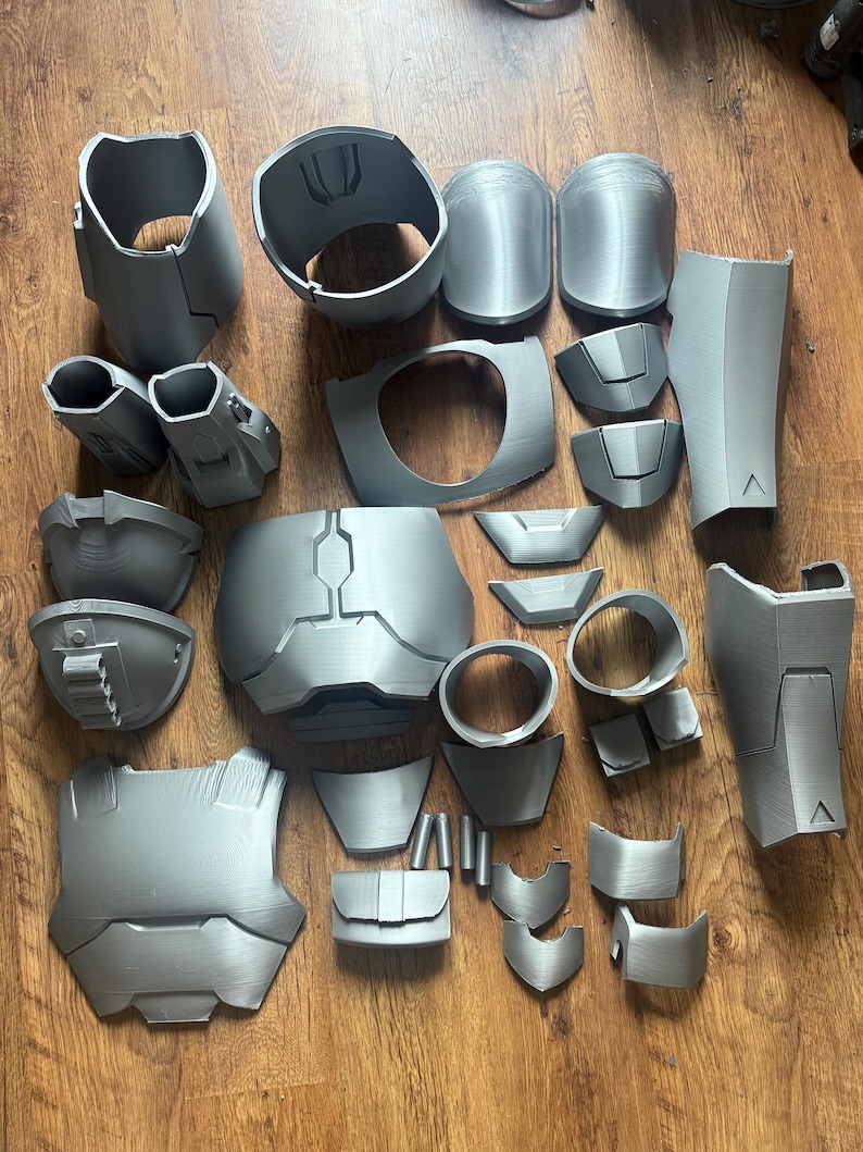 Puede incluir: Una colecci&oacute;n de piezas de armadura grises impresas en 3D, que incluyen petos, protectores de hombros y componentes de brazos. Las piezas est&aacute;n dispuestas sobre una superficie de madera, mostrando las diversas partes de un disfraz o accesorio.