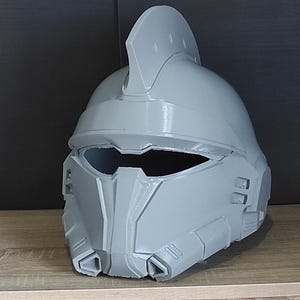 以下が含まれることがあります： 尖った紋章とバイザーが付いた灰色のプラスチック製のヘルメット。ヘルメットはテクスチャ加工された表面と詳細なデザインをしています。