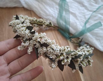 May include: A floral crown featuring small white flowers and dark green leaves. The crown is held by a hand and has a green ribbon attached. The crown is on a wooden surface.