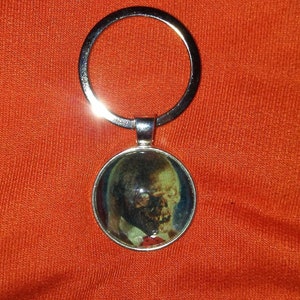 May include: A silver-colored keychain with a circular pendant featuring a zombie face. The zombie has a pale green complexion, dark eyes, and is wearing a red tie. The keychain is set against an orange background.