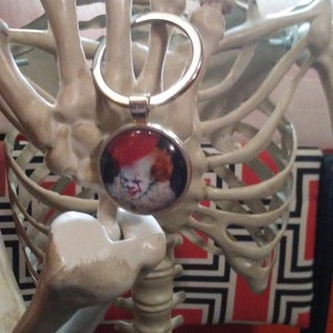 May include: A silver-colored keychain with a round pendant featuring a portrait of a clown with red hair and makeup. The keychain is held by a skeleton hand, with a rib cage in the background.