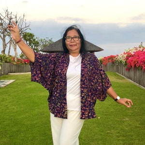 May include: A woman wearing a white top and white pants with a purple and gold patterned kimono jacket. She is standing in a grassy area with a fence and flowers in the background.