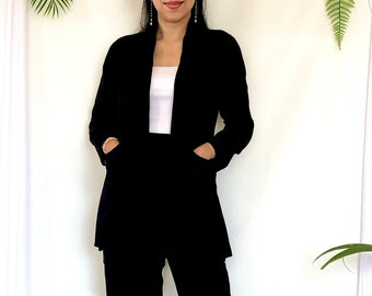 Pode incluir: Uma mulher vestindo um tailleur preto com uma camiseta branca por baixo. Ela está em pé em frente a um fundo branco com plantas verdes.