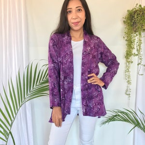 May include: A purple open-front jacket with a floral pattern. The jacket is worn over a white top and paired with white pants. The jacket's design features shades of purple and white.