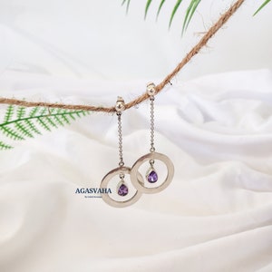 May include: A pair of silver earrings with a dangling circle and a small purple gemstone. The earrings are hanging from a rope.