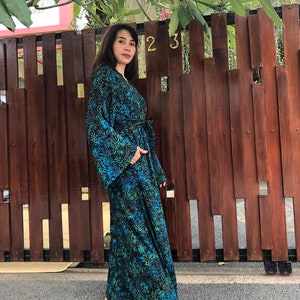 May include: A long, flowing blue and green patterned robe with a tie belt. The robe has a floral print and is worn by a person standing in front of a wooden fence.