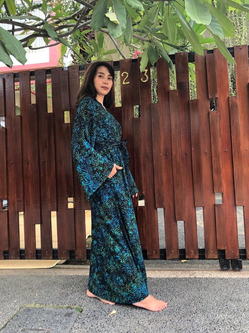 May include: A long, flowing blue and green patterned robe with a tie belt. The robe has a floral print and is worn by a person standing in front of a wooden fence.