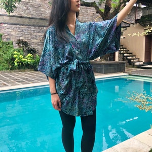 May include: A woman wearing a blue and purple floral print kimono with a black belt. She is standing by a swimming pool and wearing black leggings and black sandals.