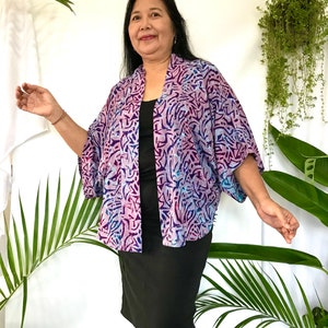 May include: A vibrant, open-front kimono in shades of purple, blue, and pink, featuring a batik-style pattern. The kimono is worn over a black dress. The image is set against a white background with green foliage.