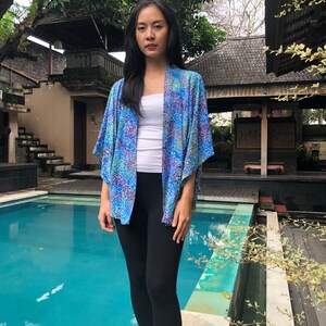 May include: A woman wearing a blue and white patterned kimono with a white tank top and black leggings. She is standing by a swimming pool.