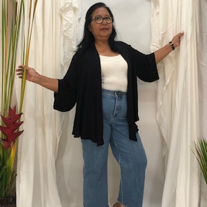 Può includere: Un cardigan nero aperto con maniche svasate, abbinato a un top bianco e jeans di denim blu chiaro. I jeans hanno un orlo grezzo. L'outfit è casual e confortevole.