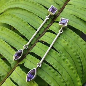 May include: A pair of silver earrings with purple gemstones. Each earring features a square gemstone at the top, a long silver bar, and a teardrop-shaped gemstone at the bottom. The earrings are displayed on a green leaf.