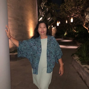 May include: A woman wearing a light green dress and a blue patterned kimono-style jacket. The jacket features a repeating leaf design. The background shows a stone wall and a pathway with hanging lights, suggesting an outdoor evening setting.