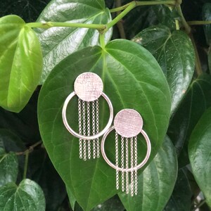 Puede incluir: Pendientes plateados con una parte superior circular texturizada y cadenas colgantes. Los pendientes se presentan sobre un fondo de hojas verdes. El diseño es moderno y minimalista, destacando el contraste entre el metal y la vegetación.